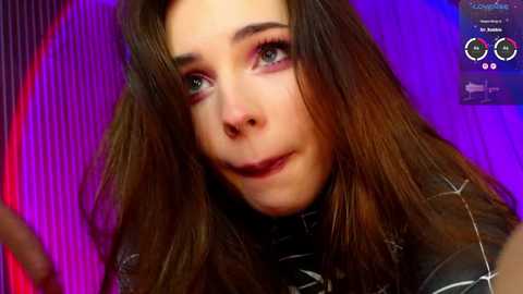 Media: Video of a young woman with long brown hair, wearing dark lipstick and black turtleneck, against a vibrant purple background. Her expression is neutral, with a hint of makeup.