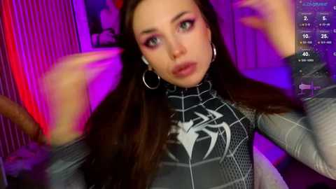Media: Video of a young woman with long brown hair, wearing a Spider-Man suit, large hoop earrings, and a surprised expression, surrounded by purple and pink lights.