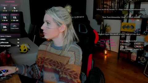 Video of a blonde woman in glasses, wearing a patterned sweater, playing a video game on a monitor, surrounded by gaming gear and a dark room.