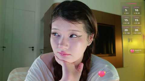 Video of a young Asian woman with fair skin, blue eyes, and long dark hair, looking contemplative. She wears a white shirt with a pink flower emblem. Background features a wooden wall and a door.