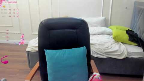 Video of a messy bedroom with a black office chair covered in a bright blue cushion, a bed with white sheets and green pillows, and scattered toys on the wooden floor.