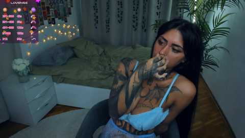 Video of a tattooed woman with dark hair, wearing a blue bra, licking her fingers in a dimly lit bedroom with a bed, potted plant, and fairy lights.