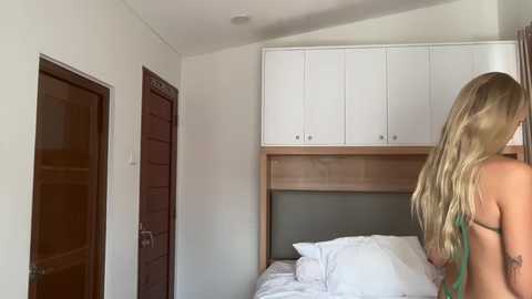 Video of a blonde woman with long, wavy hair, seen from behind, standing beside a neatly made bed in a minimalist bedroom with white walls, wooden doors, and white cabinets.