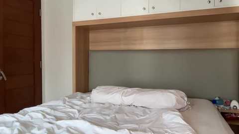Media: Video of a minimalist bedroom with a neatly made bed, white linens, and a light wooden headboard. The room has white cabinets above the headboard and a wooden door on the left.
