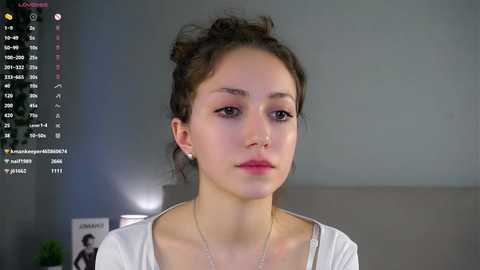 Video of a young woman with light skin and dark hair in a messy bun, wearing a white top, against a gray background.