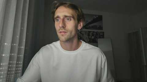 Media: A video of a Caucasian man with short brown hair and a white t-shirt, looking serious, in a dimly lit room with a white curtain and framed photos on the wall.