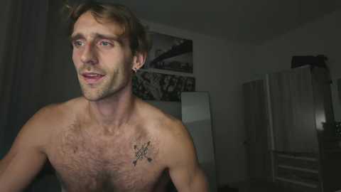Video of a shirtless, light-skinned, bearded Caucasian man with short brown hair, wearing small earrings, standing in a dimly lit room with a tattoo on his chest.
