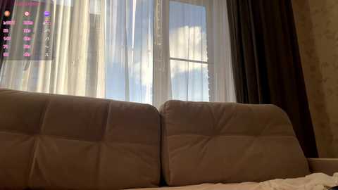 Video of a cozy, dimly lit bedroom with a plush, beige sofa in front of a large window with sheer white curtains and dark drapes. The background reveals a cloudy sky through the window, and a television screen displaying a menu is visible in the top left corner.