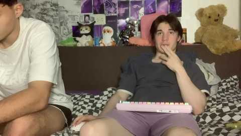 Video of two young men on a bed; one with a gaming controller, the other holding a pink keyboard, surrounded by stuffed toys and a wall collage.