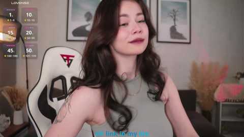 A video of a young woman with long, wavy black hair, wearing a gray tank top, seated in a white gaming chair with a red logo, in a modern living room with framed art and potted plants.