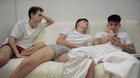 Video of three young men in white T-shirts and boxer shorts, lounging on a white couch, watching a phone, against a plain white background.
