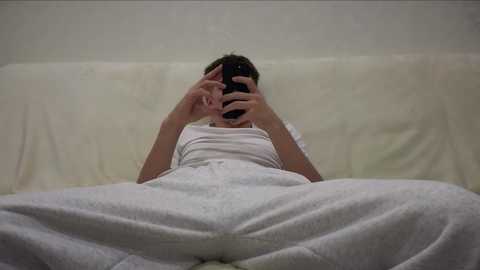 A video of a person in white pajamas lying on a white couch, covering their face with their hands, creating a sense of privacy and solitude. The background is plain and neutral, emphasizing the subject's isolation.