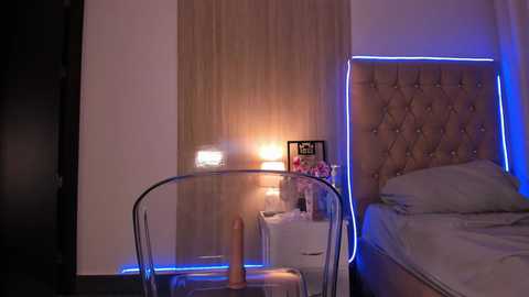 Video of a modern bedroom with a transparent chair, beige headboard, and a lit table lamp, under blue LED strip lights, creating a cozy, intimate ambiance.