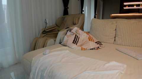 Video of a modern, plush beige sectional sofa in a well-lit room, featuring a white throw blanket and a striped pillow with orange, black, and white stripes, along with a white keyboard and a small white remote control on the surface.