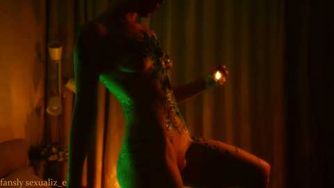 Media: Video of a nude, dark-skinned, muscular man with tattoos, illuminated by green and orange lights. He stands in a dimly lit room with green curtains, holding a lit candle.