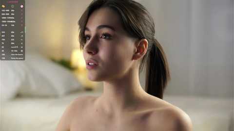 Video of a young woman with light brown skin and straight, dark brown hair tied back. She has a smooth complexion and is topless, showing a small bruise on her right shoulder. The background is softly blurred, with a hint of a bed and a lamp.