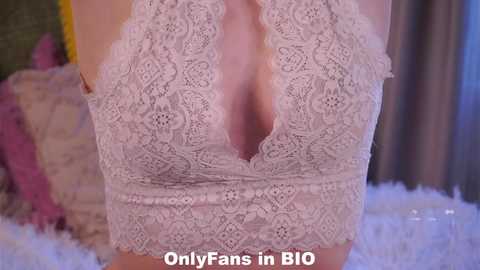 Video of a woman wearing a white lace halterneck bodysuit, revealing ample cleavage. Background includes a fluffy white rug and pastel-colored pillows. Text reads, \"OnlyFans in BIO.\