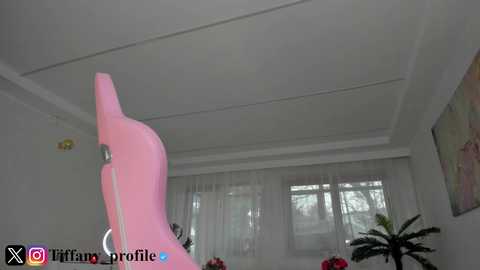 A video of a modern living room with a pink, abstract sculpture in the foreground, a white ceiling, and large windows with sheer white curtains.