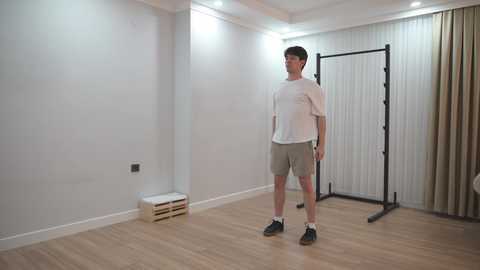 Video of a man with short hair, wearing a white T-shirt and gray shorts, standing in a minimalist, white-walled room with light wooden floors and a black exercise machine in the background.