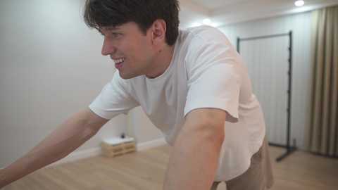 Video of a young man with short dark hair, wearing a white T-shirt and beige shorts, doing push-ups in a sparsely decorated room with wooden floors, white walls, and a black backdrop.