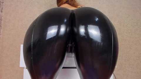 Video of a woman's bare, glossy black latex-clad buttocks, viewed from behind, on a beige textured wall. Her skin tone is light, and she's bending forward slightly.