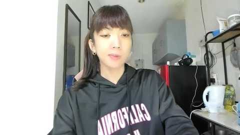 Video of an Asian woman with straight, dark hair, wearing a black hoodie with white text, standing in a cluttered, dimly lit kitchen with cabinets, a white mug, and a red fridge in the background.