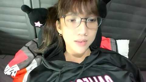 Video of an East Asian woman with glasses and cat ears, wearing a black hoodie, sitting in a car with gray seats and a red pillow.