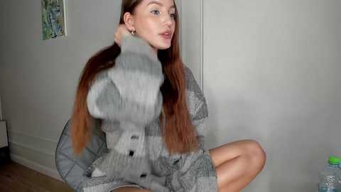 Video of a light-skinned woman with long, straight, auburn hair, wearing a grey, oversized, fur-trimmed coat, seated on a bed against a beige wall with a colorful abstract painting.
