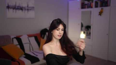 Video of a young woman with long dark hair, wearing a black off-shoulder dress, holding a lit candle, in a modern living room with white walls and colorful cushions on a sofa.