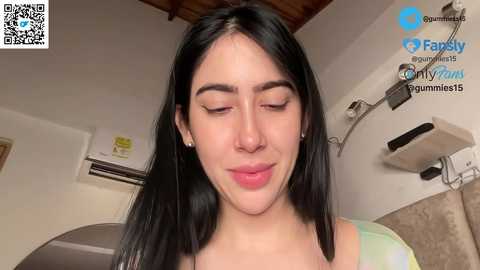 Video of a young Latina woman with long black hair, closed eyes, and a slight smile, wearing a green top, in a simple, brightly-lit room with a refrigerator and air conditioner.