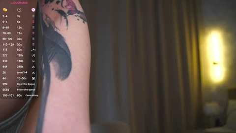 A video showing a close-up of a woman's tattooed arm, with a detailed black feather design, in a dimly lit room with yellow light.
