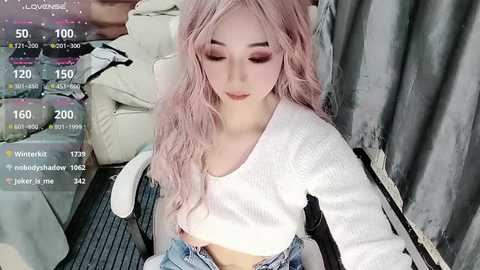 Video of a young woman with pale skin, long wavy pink hair, wearing a white cropped sweater and high-waisted jeans. She sits on a gray chair in a cozy room, surrounded by soft lighting and a cluttered background.