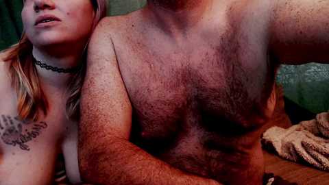 Video of a topless, muscular man with a hairy chest, and a fair-skinned woman with blonde hair and a black choker, both indoors with a textured green wall and blanket background.