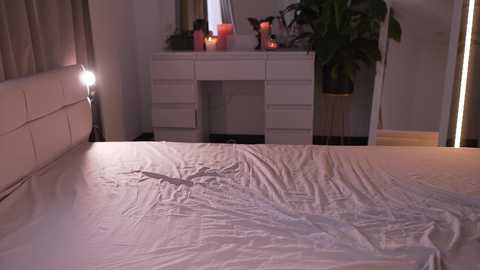 Video of a dimly-lit bedroom with a white bedspread, a white dresser holding candles and a plant, and a nightstand with a lamp.
