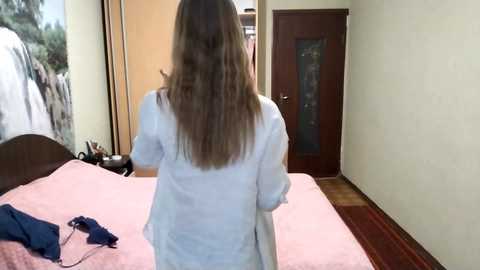 Video of a woman with long, wavy, light brown hair, standing with her back to the camera, in a bedroom with a pink bedspread, dark wooden door, and beige walls.