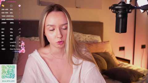 A video of a blonde woman in a white robe, looking serene, in a cozy bedroom with a bed and lamp in the background, with a webcam and text overlay.