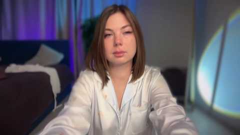 Video of a young woman with straight brown hair, wearing a light-colored jacket, sitting in a dimly lit room with a bed, curtains, and a window with a circular design in the background.