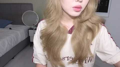 Video of a young woman with long, wavy blonde hair, wearing a white \"Warriors\" shirt, in a modern bedroom with a gray upholstered bed, white walls, and a round mirror.