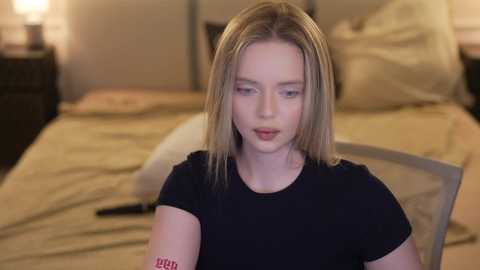 Video of a young, fair-skinned woman with straight, shoulder-length blonde hair, wearing a black t-shirt, sitting on a bed with beige linens, in a dimly lit bedroom.