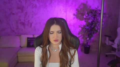 Video of a young woman with long brown hair, wearing a white blouse, seated in a black gaming chair, with a purple light background, in a cozy, dimly lit living room.