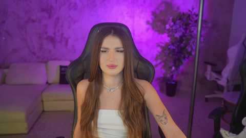 Video of a young woman with long brown hair, wearing a white strapless dress, sitting in a black gaming chair. Background features a purple-lit room with a white sectional sofa and potted plant.