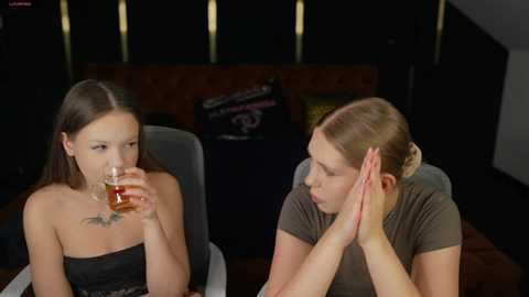 Video of two women, one with long brown hair and a black strapless top, the other with blonde hair in a bun, in a dimly lit room. They're drinking whiskey and looking at each other.