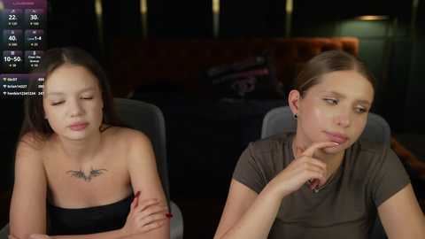 Video of two young women with fair skin, one with a bat tattoo, sitting side by side, looking contemplative. Background shows a dimly lit, modern room with a couch and lamp.