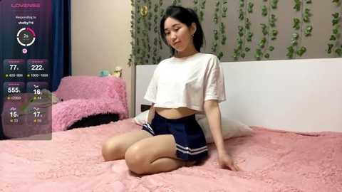 Video of an Asian woman with short black hair, sitting on a pink bed with a white wall and green ivy. She wears a white crop top and navy blue skirt, looking serene.