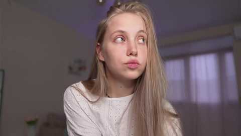 Video of a young, blonde-haired girl with fair skin and light makeup, wearing a white, textured top, looking pensively to the side. Background shows a blurred, softly lit room with a window and a few indistinct objects.