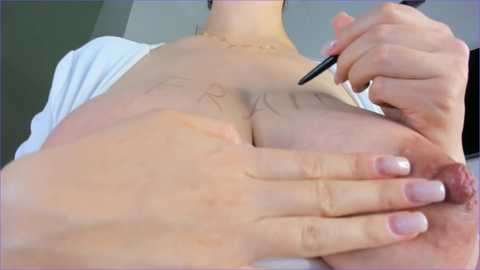 A video of a light-skinned woman with a slender build, wearing a white top, being tattooed on her chest. She has a gold necklace and a black pen in her right hand.