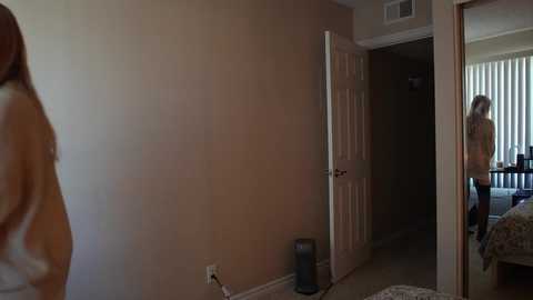 Video of a beige-walled bedroom with open door revealing another room. A woman with long, light brown hair in a white shirt stands in the doorway, while a woman in a floral dress stands in the adjacent room.