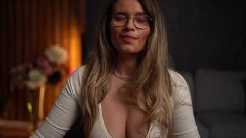 Video of a Caucasian woman with long, wavy blonde hair, wearing glasses, a low-cut white top, and a pearl necklace, sitting in a dimly-lit room with a floral arrangement in the background.