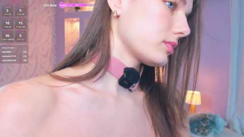 Video of a young, fair-skinned woman with long brown hair, wearing a pink choker and minimal makeup, in a softly lit, cozy bedroom.