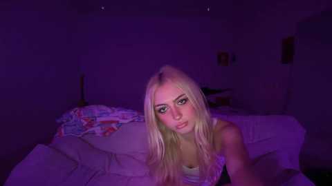 A video of a young woman with long, platinum blonde hair, wearing a sleeveless top, lying on a bed with disheveled white sheets in a dimly lit room bathed in purple light.